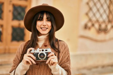 Kışlık şapkalı esmer kadın şehrin dışında klasik kamera kullanarak gülümsüyor.