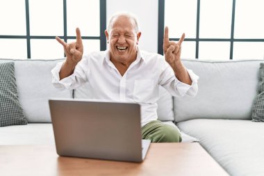 Üst sınıftaki adam evinde dizüstü bilgisayar kullanıyor. Kanepede oturmuş deli gibi bağırıyor. Elleri havada rock sembolü yapıyor. Müzik yıldızı. ağır kavram. 