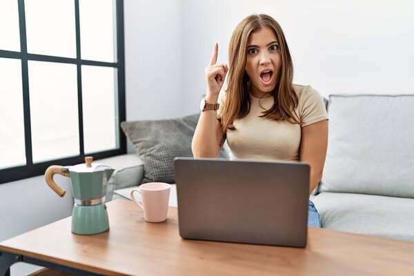 Молодая брюнетка, сидящая дома с ноутбуком, пьет чашку кофе, показывая пальцем на удачную идею. Взволнован и счастлив. номер один. 