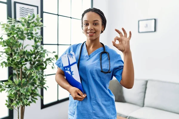 Young african american woman working wearing doctor uniform working at the clinic doing ok sign with fingers, smiling friendly gesturing excellent symbol