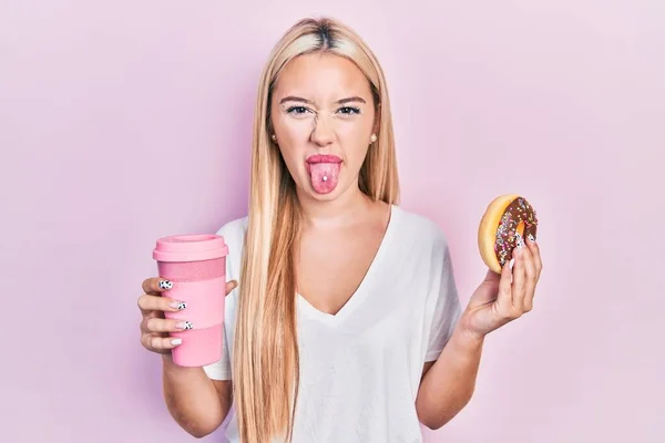 年轻的金发女郎吃着甜甜圈 喝咖啡 伸出舌头 脸上带着滑稽的表情 — 图库照片