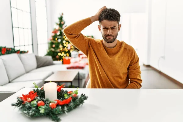 Arab young man sitting on the table by christmas tree confuse and wonder about question. uncertain with doubt, thinking with hand on head. pensive concept.