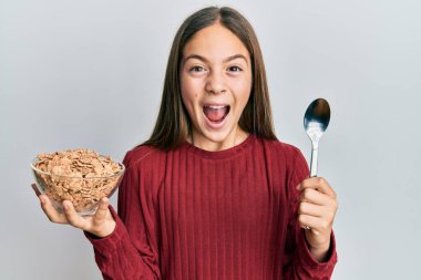 Güzel esmer küçük kız sağlıklı tahıl gevrekleri yiyor. Kaşıkla kutlama yapıyor. Başarı için hayrete düşüyor.. 