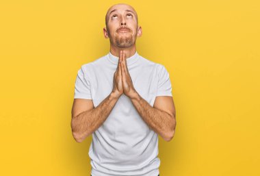 Kel adam, sakallı, günlük beyaz tişörtlü, yalvaran ve dua eden, elleri bağlı, yüzünde umut ifadesi olan çok duygusal ve endişeli bir adam. Yalvarıyor. 