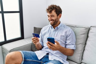 Genç İspanyol adam akıllı telefon ve kredi kartı kullanıyor. Evdeki kanepede oturuyor..