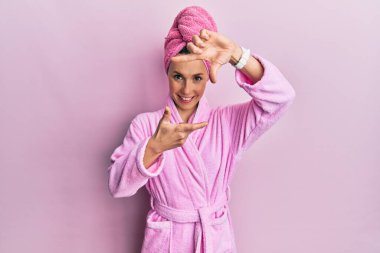 Duş başlığı ve bornoz takan genç sarışın kadın gülümsüyor elleri ve parmakları gülen yüzlü olan bir çerçeve yapıyor. yaratıcılık ve fotoğrafçılık kavramı. 