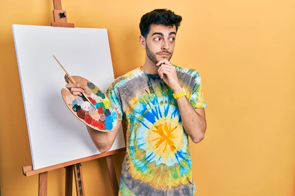 画家イーゼル スタンドによるパレットで絵を描く若いヒスパニック系の男は 質問を心配し 顎に手で心配し — ストック写真