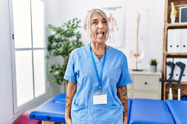 中年妇女白发苍苍 身穿理疗师制服 站在医务室里 带着滑稽的表情伸出舌头 高兴极了 情感概念 — 图库照片