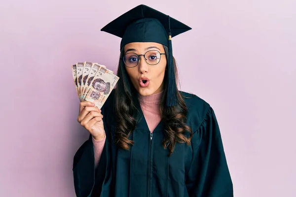 卒業制服を着た若いヒスパニック系の女性がメキシコペソを持って怖い紙幣を保持し 驚きのために口を開けて驚いて 不信の顔 — ストック写真