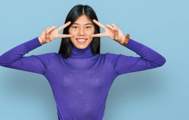 Güzel, genç Asyalı bir kadın günlük kıyafetler giyerek barış sembolü yapıyor. Yüzünde parmakları olan, neşeli bir gülümsemeyle zaferini gösteriyor. 