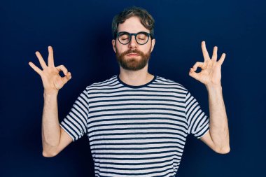 Sakallı, çizgili tişört giyen, gözlüklü, gözleri kapalı gülümseyen, parmaklarıyla meditasyon hareketi yapan beyaz bir adam. yoga konsepti. 