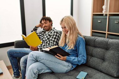 Mutlu gülümseyen genç çift evdeki kanepede oturmuş kitap okuyor..