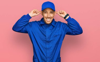 Sakallı, inşaatçı tulumu giymiş, kel bir adam. Gülümsüyor, kulakları parmaklarıyla çekiyor. Komik bir jest. deneme sorunu 