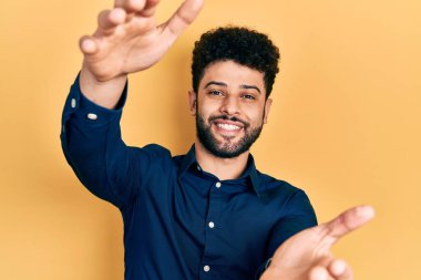 Sakallı, sıradan gömlekli genç bir adam kameraya bakıp sarılmak için kollarını açmış gülümsüyor. Mutluluğu kucaklayan neşeli bir ifade.. 