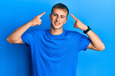 Sıradan mavi tişört giyen genç beyaz adam iki eliyle başını gösteren gülümseme, harika bir fikir ya da düşünce, iyi hafıza 