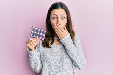 Genç esmer kadın elinde haplarla ağzını eliyle kapatıyor, şokta ve hata yapmaktan korkuyor. şaşırmış ifade 