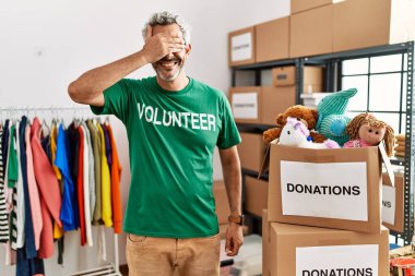 Bağışta gönüllü tişört giyen orta yaşlı İspanyol adam gülümseyerek ayakta duruyor ve elleri yüzünde şaşkınlıktan gözleri kapatıyordu. kör kavram. 