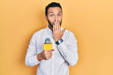 Gazeteci sakallı İspanyol bir adam mikrofon tutarak ağzını eliyle kapatıyor, şok geçiriyor ve hata yapmaktan korkuyor. şaşırmış ifade 