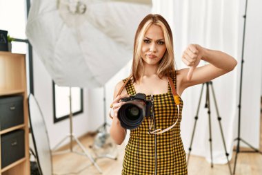 Kafkasyalı genç fotoğrafçı kız elinde kızgın suratlı profesyonel kamerayla fotoğraf stüdyosunda. Negatif işaretler baş parmakları aşağıda, reddedilme konsepti ise reddedilmiş. 