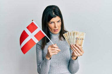 Genç İspanyol kadın elinde Danimarka bayrağı ve krone banknotlarıyla şaşkın, şüpheci ve alaycı bakıyor, ağzı açık kalmış. 