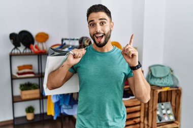 Alışveriş torbaları tutan sakallı yakışıklı genç adam başarılı bir fikirle parmakla işaret ediyor. Heyecanlı ve mutlu. Bir numara.. 