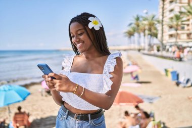Genç Afrikalı Amerikalı kız sahilde akıllı telefon kullanarak mutlu bir şekilde gülümsüyor..