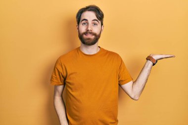 Sakallı, sıradan sarı tişörtlü, neşeli gülümseyen, eli avucunda kameraya bakan beyaz bir adam.. 