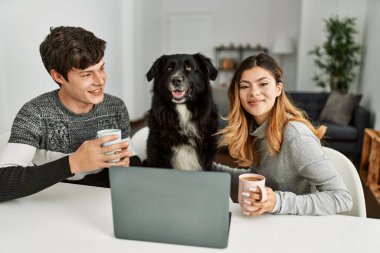 Genç beyaz çift dizüstü bilgisayar kullanıyor ve kahve içiyor. Evde köpeklerle birlikte masada oturuyorlar..