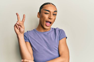 Makyaj yapan, uzun saçlı, sıradan tişörtlü, gülümseyen, kameraya göz kırpan ve zafer işareti yapan İspanyol bir adam. İki numara.. 