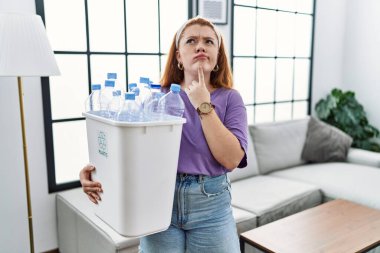 Genç kızıl saçlı kadın elinde plastik şişelerle çöp sepetini tutarken parmakları çenesinde şüpheyi düşünüp kafasını kaldırıyor. 