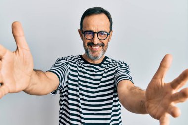 Orta yaşlı İspanyol bir adam günlük kıyafetler ve gözlüklerle kameraya bakıp kucaklaşmak için kollarını açmış gülümsüyor. Mutluluğu kucaklayan neşeli bir ifade.. 