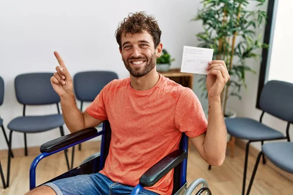 年轻英俊的男子拿着漂亮的记录卡坐在轮椅上 高兴地微笑着 手指手画脚地指向旁边 — 图库照片