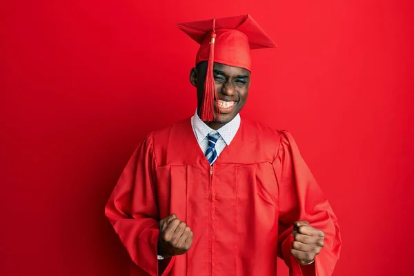 身穿毕业典礼帽和礼服的年轻的非洲裔美国人兴奋地举起双臂 闭上双眼 微笑着庆祝胜利 获奖者概念 — 图库照片