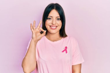Tişörtüne pembe kanser kurdelesi takmış genç İspanyol kadın gülümsüyor ve el ve parmaklarıyla 