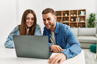 Genç beyaz çift evde bilgisayar kullanarak çalışıyor. Olumlu ve mutlu görünüyorlar. Dişlerini gösteren bir gülümsemeyle duruyorlar. 