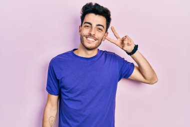 Gündelik tişört giyen genç İspanyol adam barış sembolü yapıyor. Yüzünde parmaklar, gülümseyerek zaferi gösteriyor. 