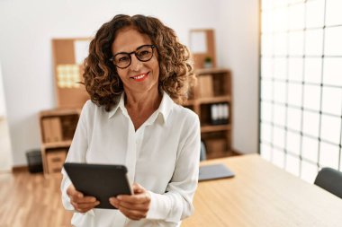 Orta yaşlı İspanyol kadın, ofiste dokunmatik ped kullanarak çalışırken kendine güvenerek gülümsüyor.