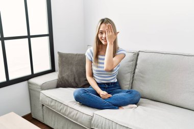 Asyalı genç bir kadın evdeki kanepede oturuyor bir gözünü eliyle kapatıyor, yüzünde kendine güvenen bir gülümseme ve sürpriz bir duygu var.. 