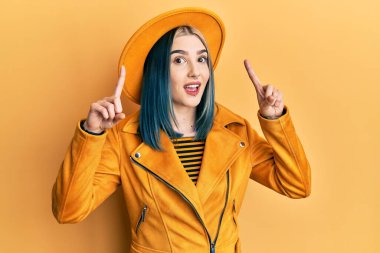 Sarı şapka ve deri ceket giyen genç ve modern bir kız. Şaşırmış, şaşırmış, parmaklarıyla işaret ediyor ve kollarını kaldırıyordu.. 