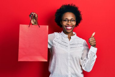 Afro-Amerikalı genç bir kadın alışveriş torbaları tutuyor mutlu ve pozitif gülümsüyor, başparmağıyla mükemmel ve onay işareti yapıyor. 