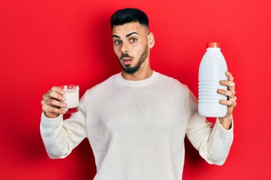 Sakallı, elinde bir bardak süt ve plastik şişeyle şaşkın bakışlı, şüpheci ve alaycı bakışlı, ağzı açık bir şekilde şaşırmış genç bir İspanyol. 