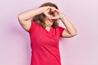 Orta yaşlı beyaz kadın gündelik giysiler giyip el ve parmaklarıyla kalp şeklinde tabelalardan bakıp gülümsüyor. 