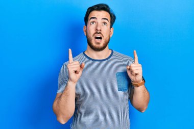 Sıradan çizgili tişört giyen yakışıklı beyaz adam şaşırmış ve şaşırmış görünüyor ve parmaklarıyla işaret ediyor ve kollarını kaldırıyor.. 