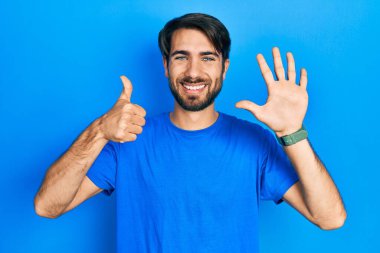 Latin asıllı genç adam gündelik giysiler giyip altı numaralı parmaklarıyla işaret ederken kendinden emin ve mutlu bir şekilde gülümsüyor.. 