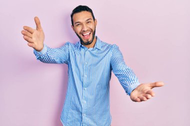 Sakallı, sıradan mavi tişörtlü, kucaklaşmak için kollarını açıp kameraya gülümseyen İspanyol adam. Mutluluğu kucaklayan neşeli bir ifade.. 