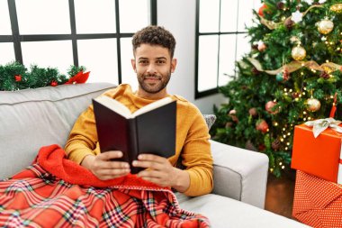 Genç Arap adam kitap okuyor. Evdeki Noel ağacının yanındaki kanepede yatıyor..