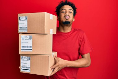 Sakallı, teslimat paketleri taşıyan, komik suratlı, yanak şişiren genç Afro-Amerikalı adam. Ağzı havayla şişer, havayı yakalar.. 