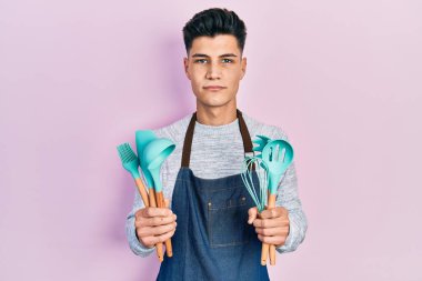 Profesyonel fırıncı önlüğü giyen genç İspanyol adam elinde yemek aletleri tutarken yüzünde ciddi bir ifadeyle rahatladı. Basit ve doğal bir şekilde kameraya bakıyorsun.. 