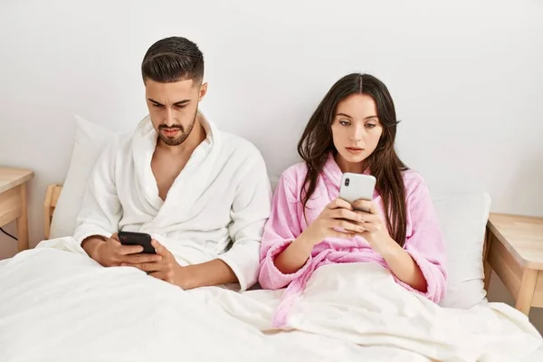 Yatakta Uzanmış Akıllı Telefon Kullanan Ciddi Yüzlü Genç Spanyol Çift — Stok fotoğraf