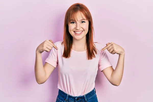 顔に笑顔で自信を持って見えるカジュアルなピンクのTシャツを着て若い女性をRedhead 誇りと幸せ指で自分自身を指して — ストック写真